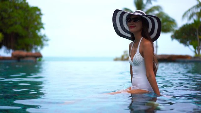 Pretty Asian girl in a one-piece white bathing suit, sunglasses and floppy straw hat sits in the shallow water of a resort pool sunbathing.