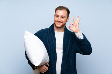 Blonde man in pajamas over blue wall showing ok sign with fingers