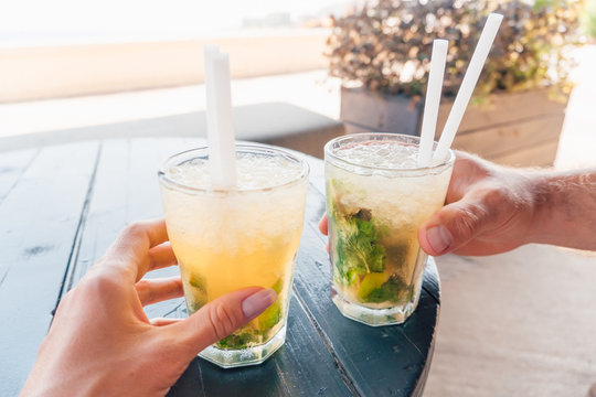Close-up of two mojito cocktails on the table - POV image of male and female hands holding glasses, people (couple) sitting in a bar