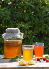 Kombucha in a glass jar and two glasses with a drink with lemon and raspberry in the foreground, in the summer garden, on a wooden table. Fermented drink.