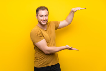 Blonde man over isolated yellow wall holding copyspace to insert an ad