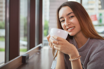 Close up of a lovely young woman smiling to the camera, holding cup of delicious fresh coffee, copy space. Happy woman enjoying morning coffee