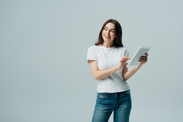 happy girl in white t-shirt holding digital tablet and looking away isolated on grey