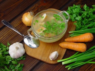 Soup with spoon on wooden cutting board over wooden table. There are onion, pepper, chili pepper, parsley, garlic on the table.