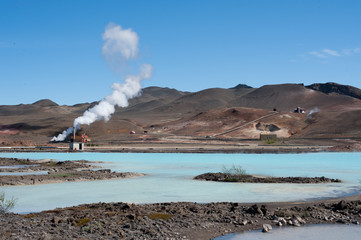  Steam comes from a geo thermal power station.Pipelines and volcanic landscape are visible with turquoise water in foreground - Image
