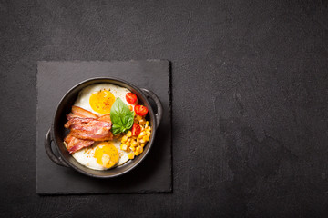 Two fried eggs with bacon, tomatoes and corn in a black pan on a dark background Top view with copy space for your text