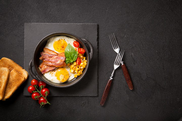 Two fried eggs with bacon, tomatoes and corn in a black pan on a dark background Top view with copy space for your text