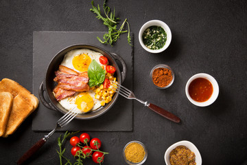Two fried eggs with bacon, tomatoes and corn in a black pan on a dark background Top view with copy space for your text