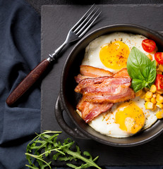 Two fried eggs with bacon, tomatoes and corn in a black pan on a dark background Top view with copy space for your text