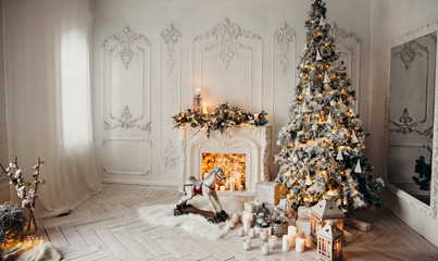 classical luxury interior of a white room with Christmas tree with garland, decorated fireplace, rocking horse, gifts for new year