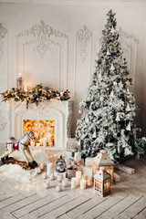 classical luxury interior of a white room with Christmas tree with garland, decorated fireplace, rocking horse, gifts for new year