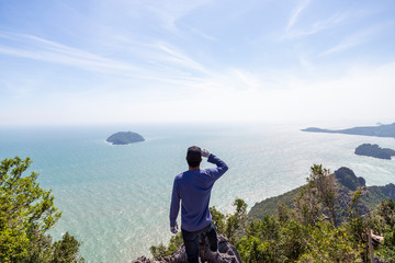 Men in blue shirts are looking at the beauty of the sea on the hilltop with blue sky. Men are looking at the success of climbing.