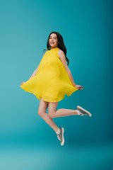 full length view of happy girl in yellow dress jumping on turquoise background