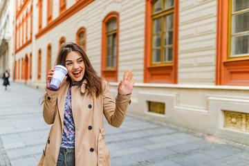 Fototapeta premium Coffee on the go. Beautiful young woman holding coffee cup and smiling while walking along the street. Young beautiful brunette drinking coffee walking around the city.