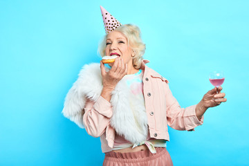 glamour old lady in party hat holding a glass of wine and eating yummy cake. isolated blue background. studio shot. people, happiness