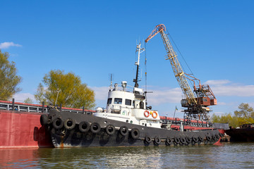 Fototapeta premium Tugboat with row of black tires used as boat bumpers in harbor near port crane