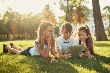 three smiling school-age friends are lying on the grass and looking at the tablet