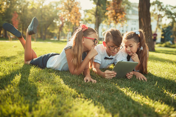 three smiling school-age friends are lying on the grass and looking at the tablet - obrazy, fototapety, plakaty