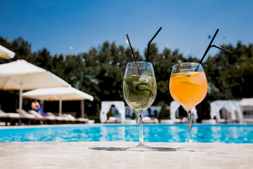 Two glasses with a cocktail stand on the edge of the swimming pool next to lounge zone on the open air on a sunny summer day
