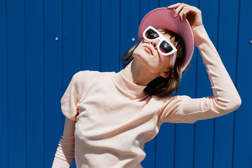 Stylish girl in a gently pink dress, white sunglasses and pink visor poses against a bright blue metal wall outside on a sunny day