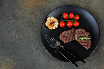 Grilled beef steak on a black plate. Grilled garlic, cherry tomatoes and cutlery in a plate. View from above. Close-up. Dark background. Free space for text.