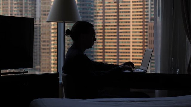 Man sitting with portable computer against window, push backspace button to delete message. Dark silhouette, dim natural light from outside. Large residential building seen blurred outside
