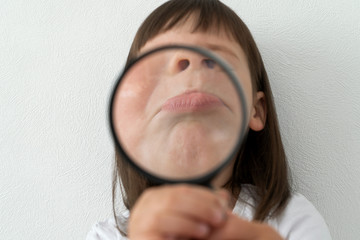 A child is looking through a magnifying glass. A large mouth of a child behind a magnifying glass. Little girl shows her mouth. Little girl studies her face