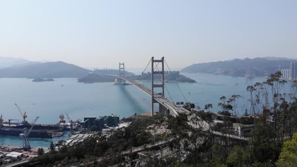 Camera fly towards Tsing Yi bridge from green hill, showing interchange in front of large construction and lengthwise perspective of the bridge. Big sea channel and islands seen ahead in light mist