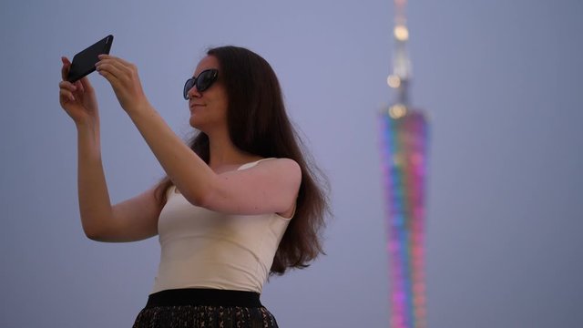 Tourist woman wear dark sunglasses take selfie at evening time, blurred Canton Tower silhouette seen on background. Twilight time
