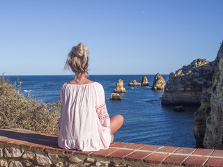 A blond lady in white shirt seen from behind sits high up and watches the colourful cliffs off Ponta da Piedade headland and cliffs rising from the turquoise waters of the sea.
