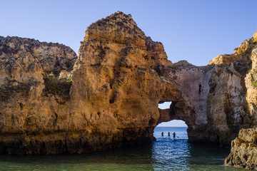 Three paddle boarders under a sandstone arch on their way from open ocean to a bay. Early morning sun makes the scene colourful. Blue sky and turquoise sea water