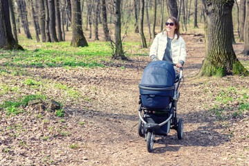 Beautiful young adult woman walking with baby in stroller through forest or park on bright sunny day. Healthy lifestyle and children healthcare. Happy childhood and parenthood