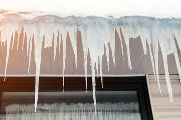 Big transparent blue icicles hanged on roof of building with window on background. Winter or spring melting seasonal danger
