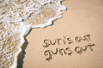 Sun's Out Guns Out summer beach holiday message handwritten on smooth sand with incoming wave