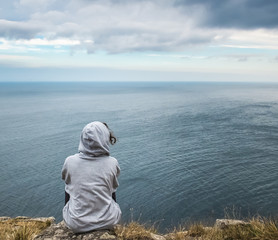 Back view of a sad, lonely little woman with a hud looking to the horizon from the cliffs of...