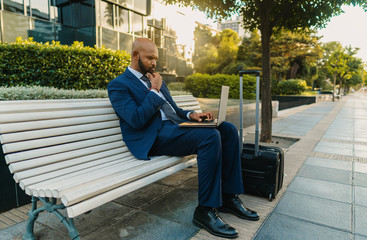 Indian businessman holding laptop notebook wearing blue suit near office. Freelance business work concept. His suitcase is near bench