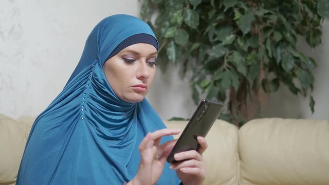 beautiful muslim woman in blue hijab using a mobile phone sitting on a sofa