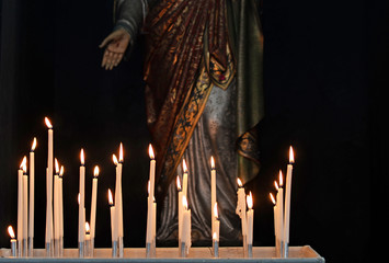 Burning white devotion candles in church from believers to pray and ask for help in front of religious sculpture of Jezus with stigmata on hand and ornate robe © annavee