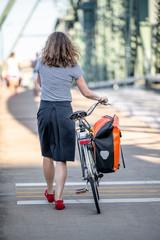 Woman walks with a bicycle with fastened bag on a footpath on a bridge