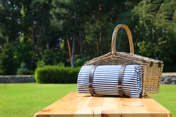 Picnic basket with blanket on wooden table in green park