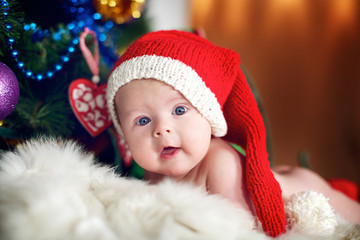 Little baby in red cap of Santa Claus celebrates Christmas. Christmas photo of infant in red cap. New Year's holidays and Christmas tree.