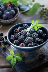 Ripe blackberries are harvested in a ceramic cup. Green leaves around and scattered berries. Wood background