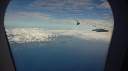 Fototapeta premium Aircraft porthole with blue sea or ocean and tropical islands. Looking through window aircraft during flight. Airplane wing through the porthole. Looking through window aircraft during flight