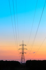 Large metal anchor the tower to the backbone transmission line is on the horizon over the dark line of trees of the forest with sagging wires on the gradient background of the colorful sky at sunset.