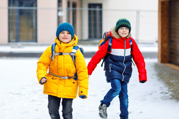 Two little kids boys of elementary class walking to school during snowfall. Happy children having fun and playing with first snow. Siblings and best friends with backpack in colorful winter clothes.