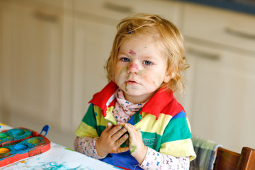 Cute adorable baby girl learning painting with water colors. Little toddler child drawing at home, using colorful brushes. Healthy happy daughter experimenting with colors, water at home or nursery.