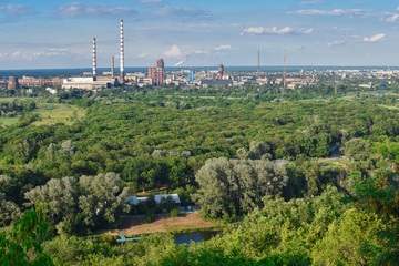 The smoking chimneys of a plant against a background of green trees, a blue river and a blue sky.