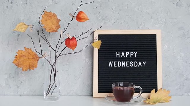 Happy Wednesday text on black letter board and bouquet of branches with yellow leaves on clothespins in vase on table agains stone gray wall. Concept Hello autumn Wednesday.