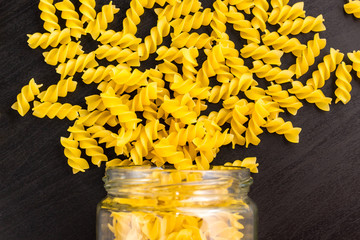 Glass jar filled with dry pasta spiral fusilli and scattered around on black background