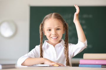 Cute little girl raising hand during lesson in classroom